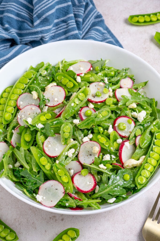 A white bowl filled with sugar snap pea salad with arugula, radishes, and feta.