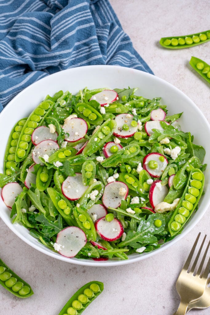 A white bowl filled with sugar snap pea salad with arugula, radishes, and feta.
