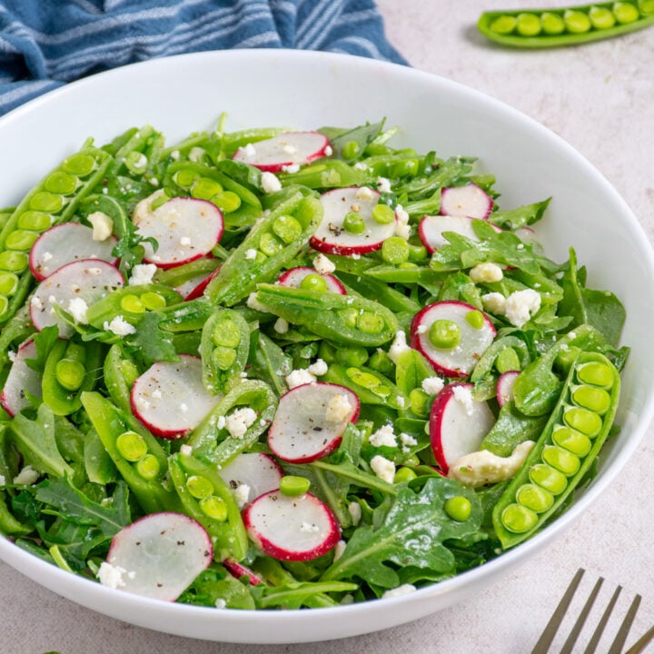 A white bowl filled with sugar snap pea salad with arugula, radishes, and feta.