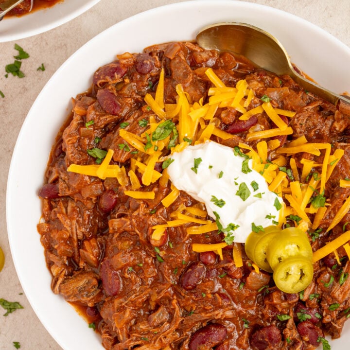 Overhead voew of a white bowl with chuck roast chili garnished with cheddar cheese, sour cream, and jalapeno slices.