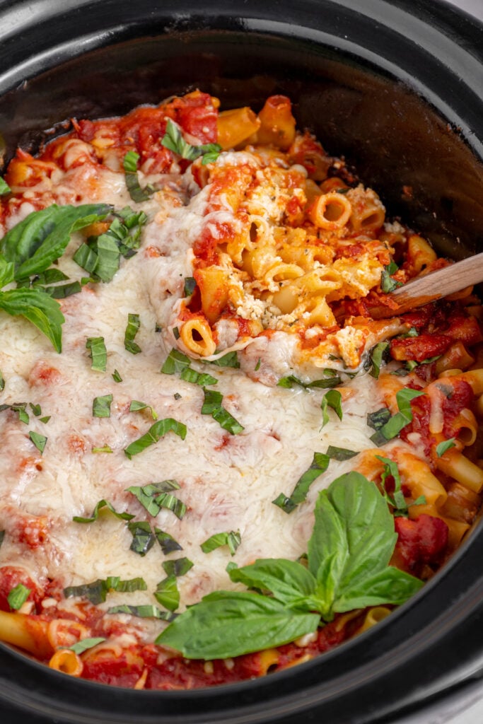 A black crockpot with tender crockpot ziti with a wooden spoon digging a scoop to reveal the pasta.
