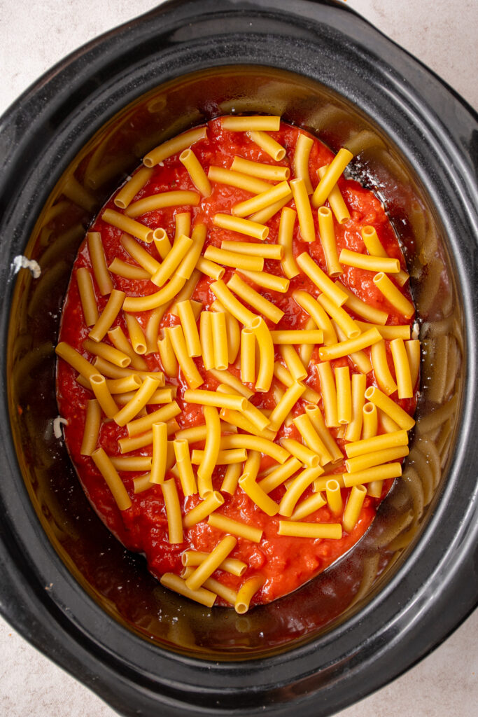 Adding uncooked ziti noodles to the crockpot over a layer of sauce.