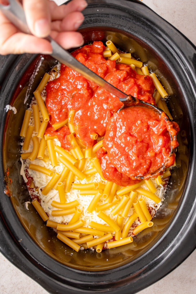 A ladle adding tomato sauce to a crockpot over top ziti noodles.