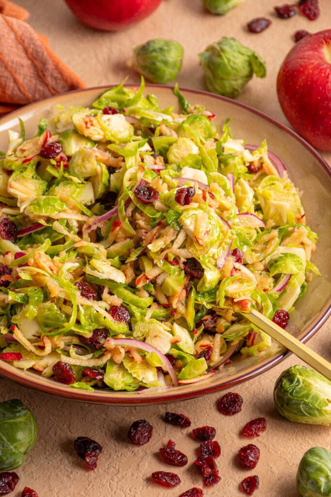 A serving fork in a brown bowl filled with Shredded Brussel Sprout Apple Salad with dried cranberries and red onions.