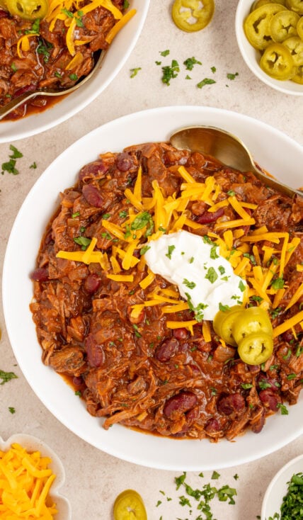 A white bowl filled with chuck roast chili garnished with cheddar cheese, sour cream, and jalapeño slices.