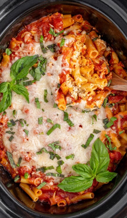A black crockpot with baked crockpot ziti with a serving spoon digging in to reveal tender pasta.