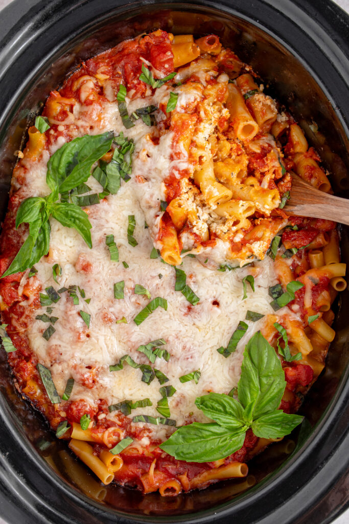 A black crockpot with baked crockpot ziti with a serving spoon digging in to reveal tender pasta.