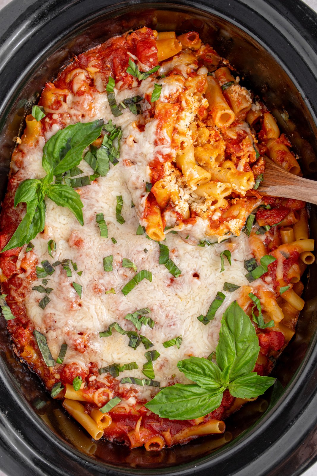 A black crockpot with baked crockpot ziti with a serving spoon digging in to reveal tender pasta.