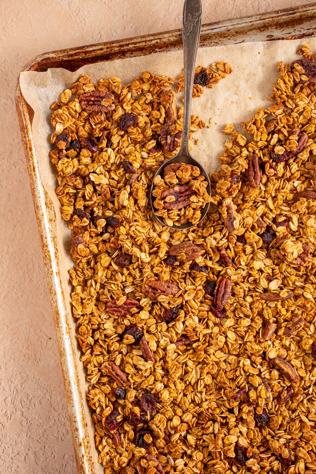 Sheet pan with parchment paper filled with pumpkin spice granola with dried cranberries and pecans with a spoon scooping some granola.
