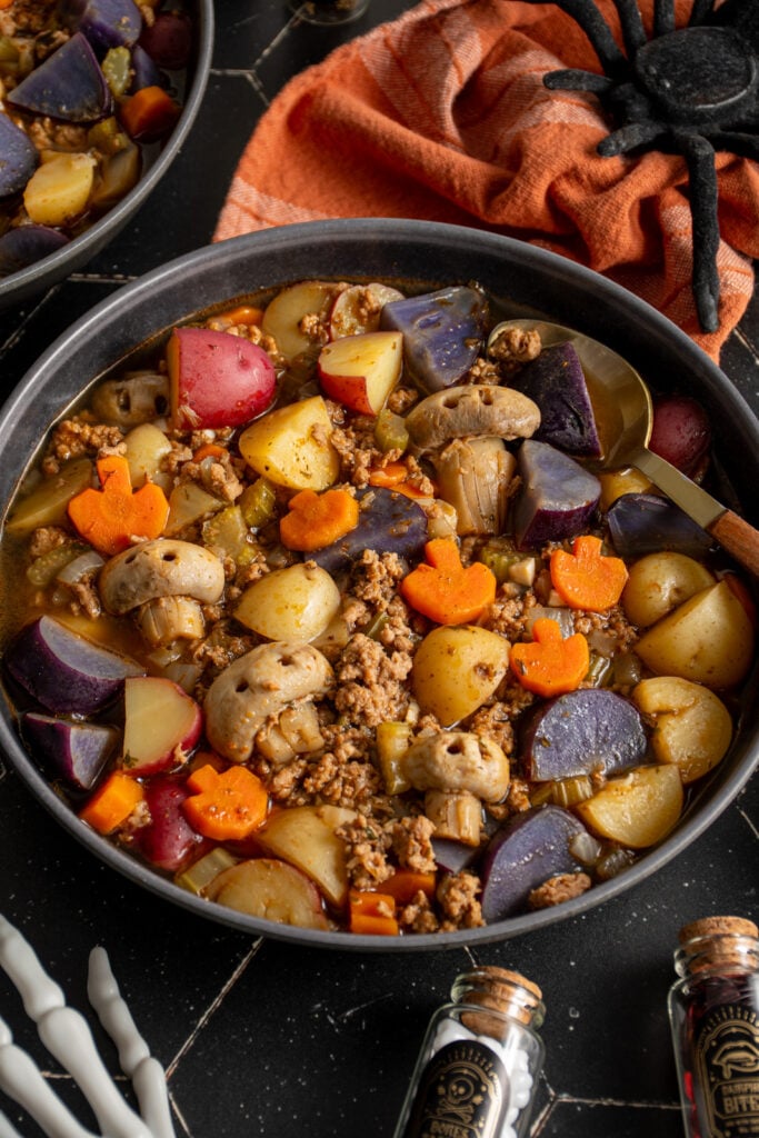 Black bowls against a black background filled with halloween soup with pumpkin shaped carrots and skull shaped mushrooms along with purple potatoes and ground turkey.