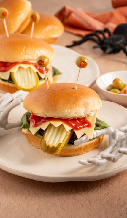 A halloween monster burger with a pickle tongue, cheese teeth, bloody ketchup, and olive eyeballs on a plate.