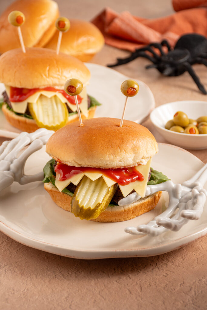 A halloween monster burger with a pickle tongue, cheese teeth, bloody ketchup, and olive eyeballs on a plate.