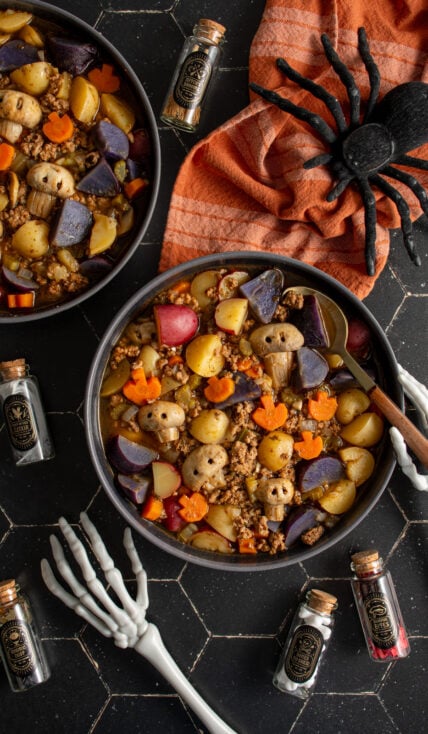 Black bowls against a black background filled with halloween soup with pumpkin shaped carrots and skull shaped mushrooms along with purple potatoes and ground turkey.