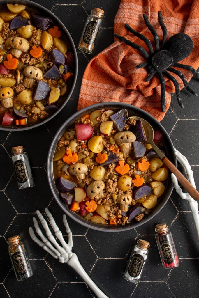 Black bowls against a black background filled with halloween soup with pumpkin shaped carrots and skull shaped mushrooms along with purple potatoes and ground turkey.