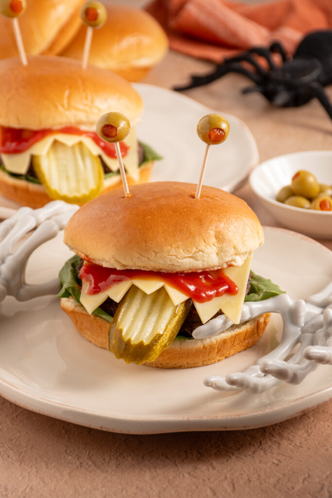 A halloween monster burger with a pickle tongue, cheese teeth, bloody ketchup, and olive eyeballs on a plate.