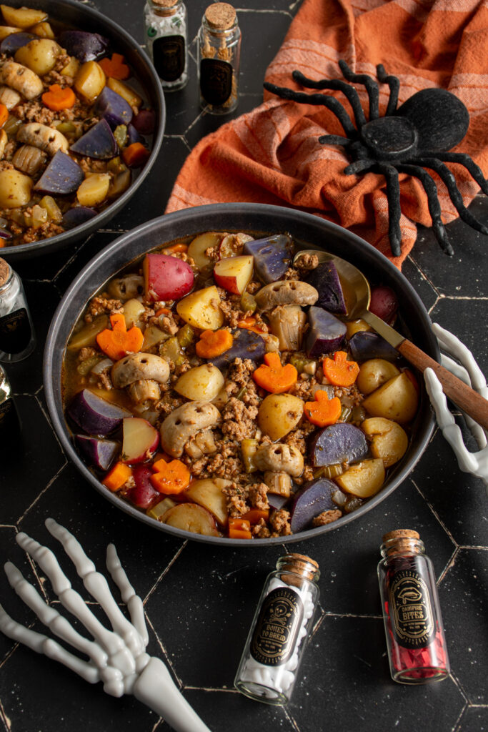 Black bowls against a black background filled with halloween soup with pumpkin shaped carrots and skull shaped mushrooms along with purple potatoes and ground turkey.