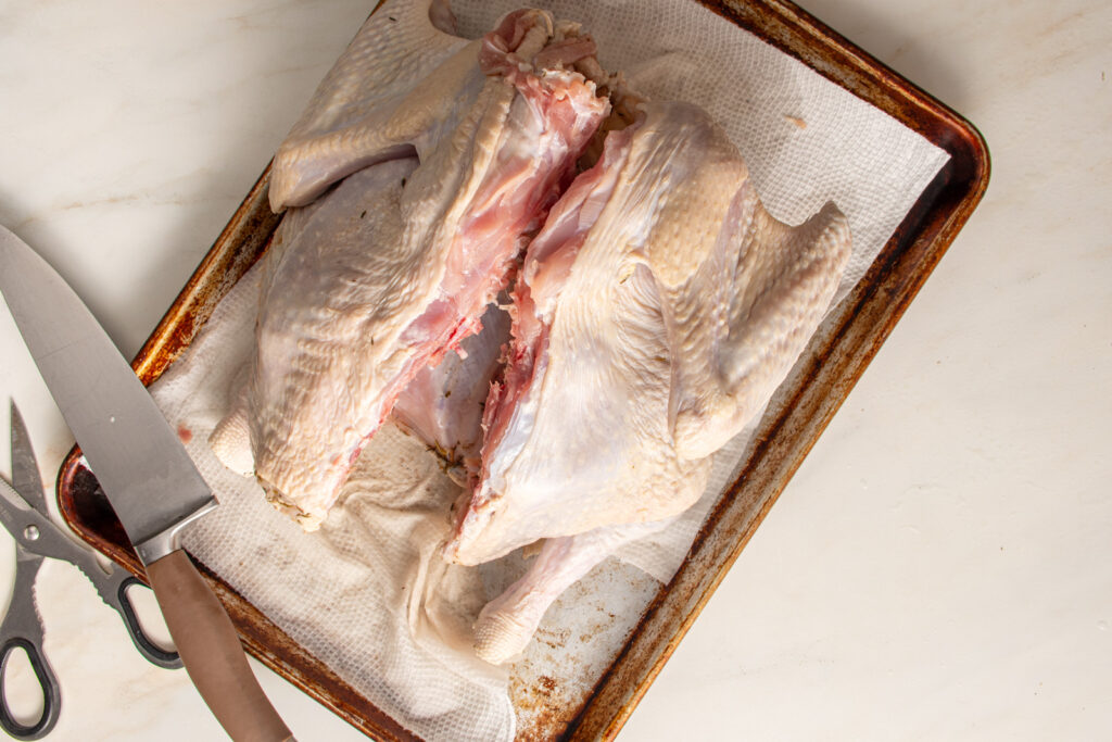 A raw turkey on a sheet pan with a cut all along one side of the backbone.