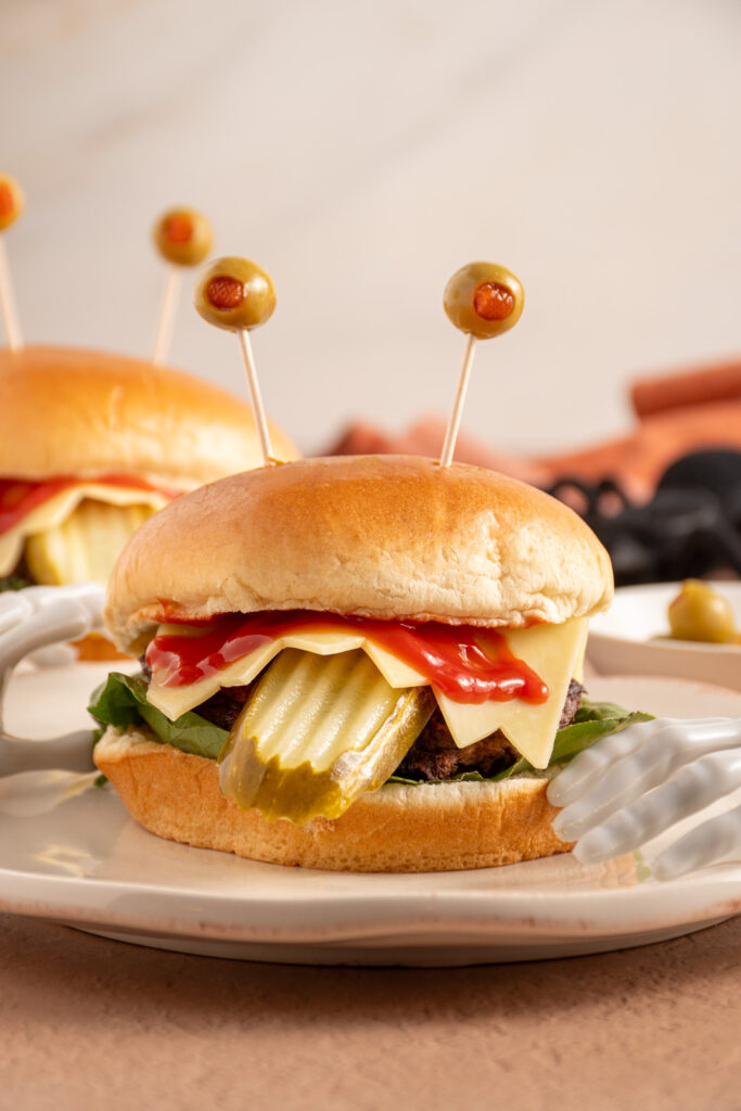 A halloween monster burger with a pickle tongue, cheese teeth, bloody ketchup, and olive eyeballs on a plate.