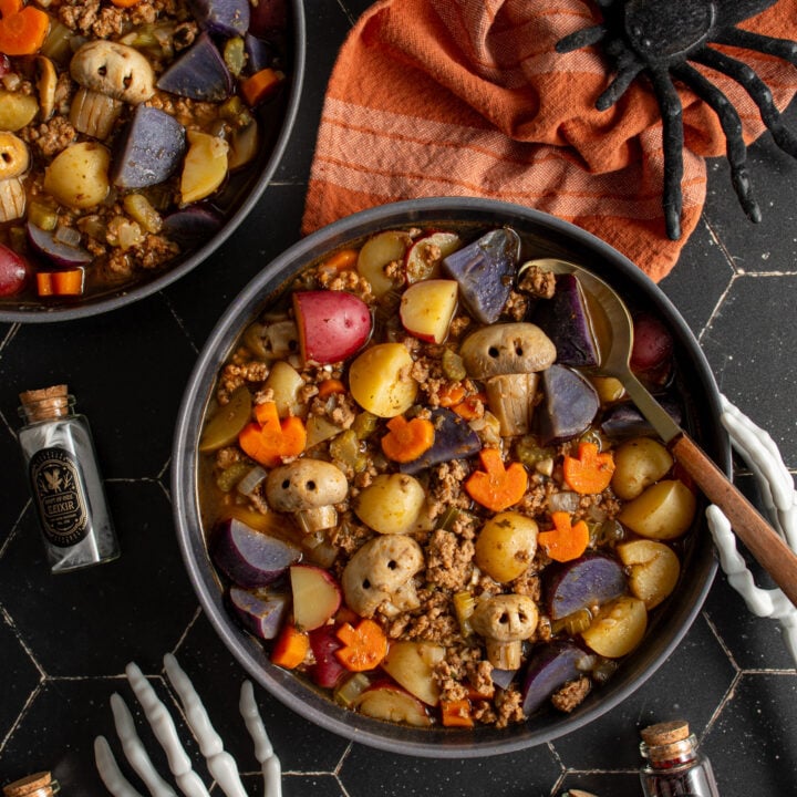 Black bowls against a black background filled with halloween soup with pumpkin shaped carrots and skull shaped mushrooms along with purple potatoes and ground turkey.