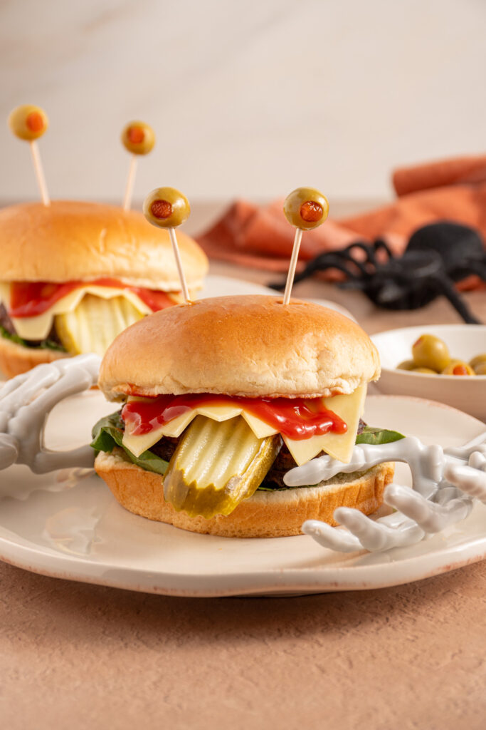 A halloween monster hamburger on a plate with cheese teeth, a pickle tongue, and olive eyeballs!