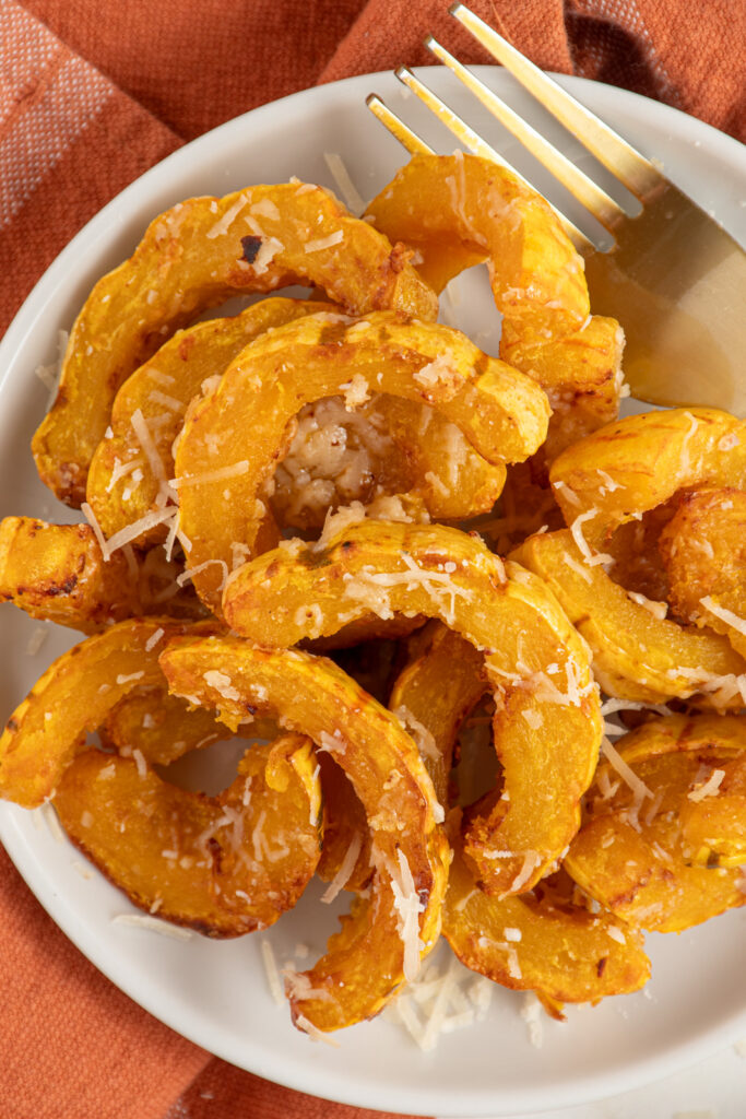 Delicata squash cooked golden brown with melted parmesan cheese on a white plate next to a fork.