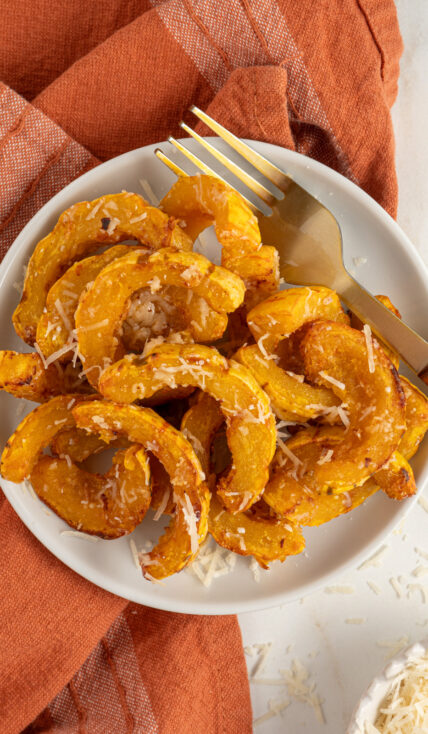 Delicata squash cooked golden brown with melted parmesan cheese on a white plate next to a fork.