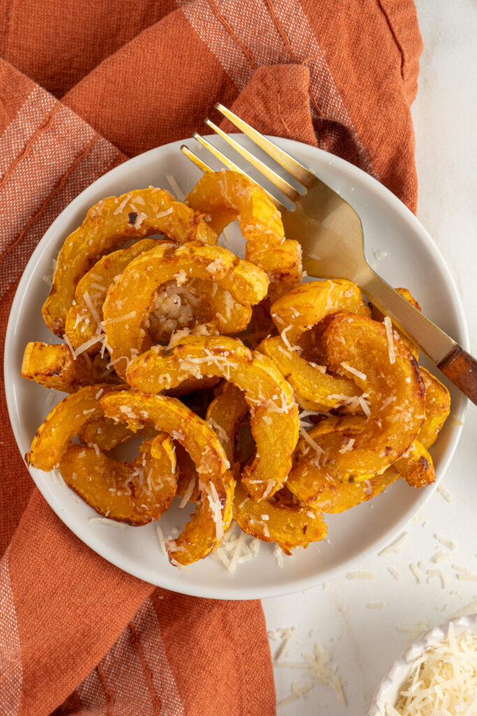 Delicata squash cooked golden brown with melted parmesan cheese on a white plate next to a fork.