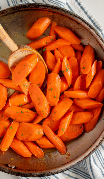 A skillet with honey glazed carrots with a wood spoon and garnished with a bit of fresh parsley.