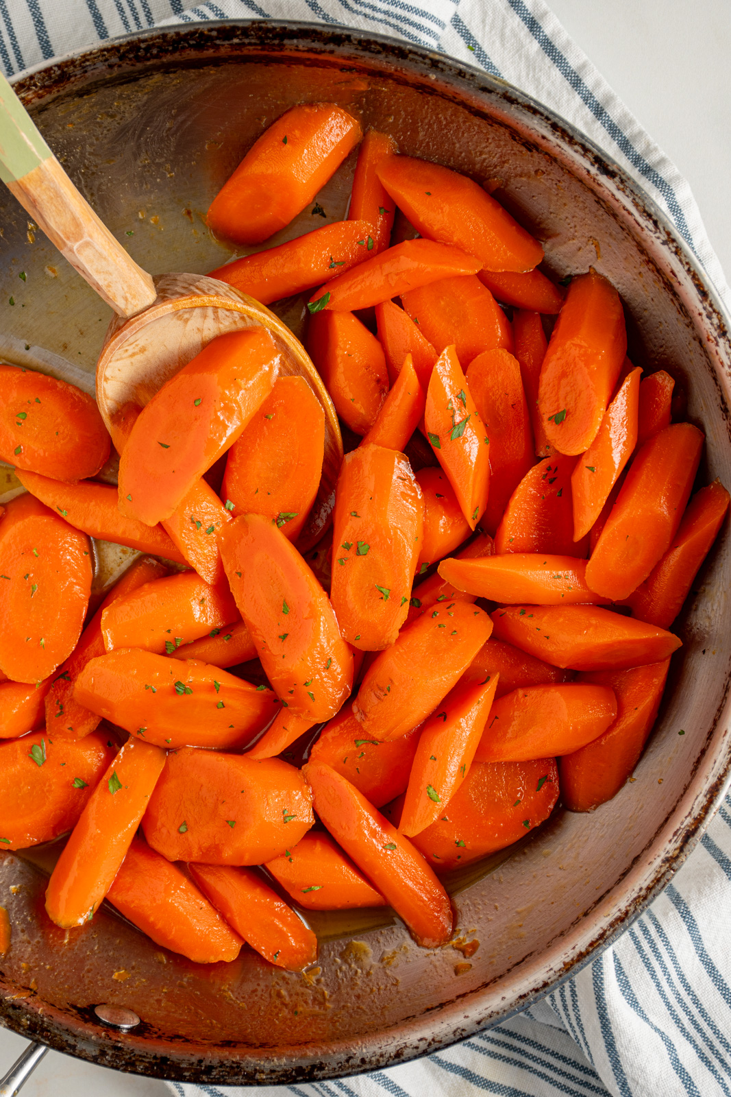 A skillet with honey glazed carrots with a wood spoon and garnished with a bit of fresh parsley.