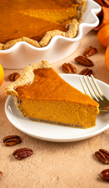 A slice of pumpkin pie with pecan crust on a small white plate surrounded by mini pumpkins and pecan halves.