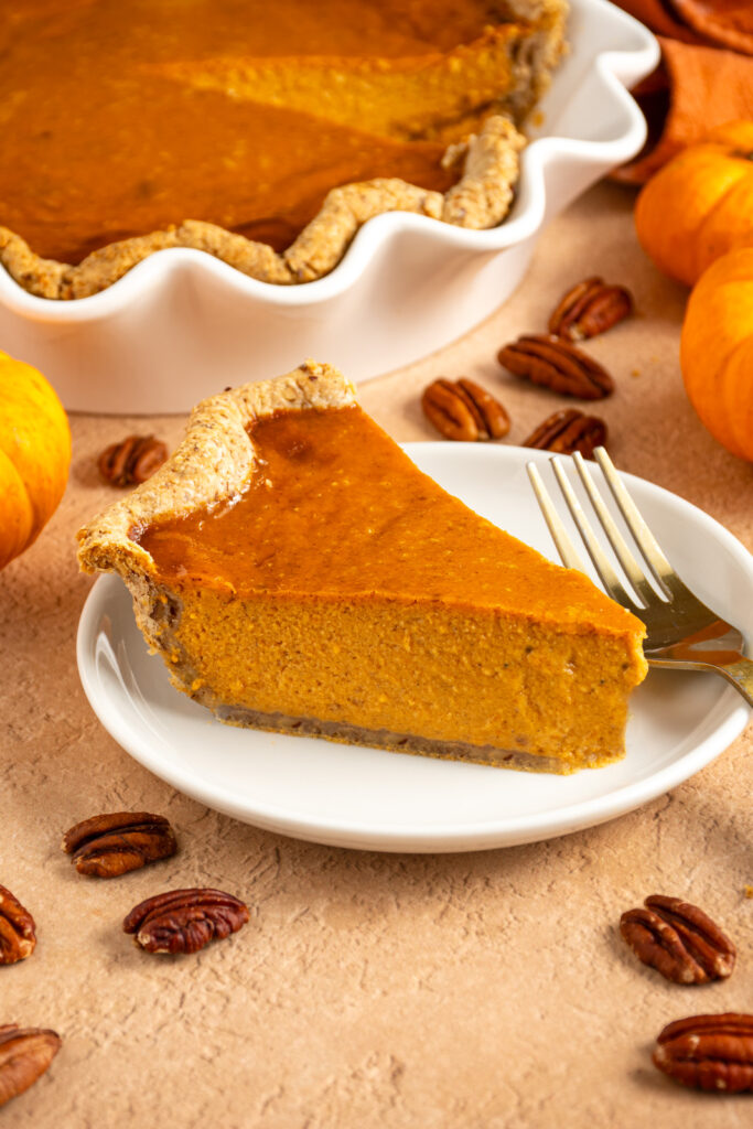 A slice of pumpkin pie with pecan crust on a small white plate surrounded by mini pumpkins and pecan halves.