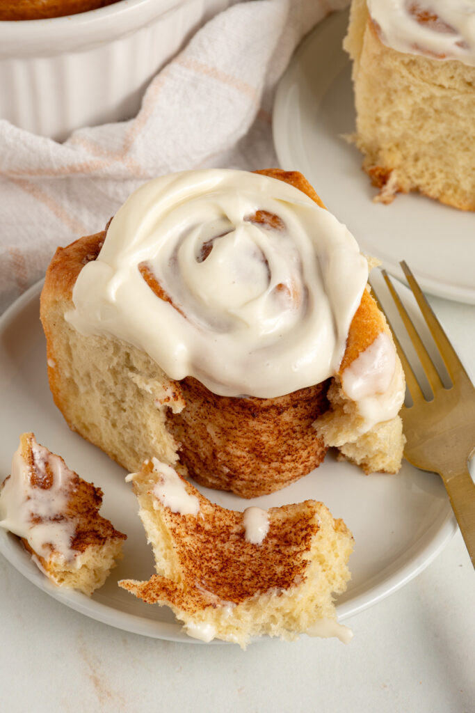 A fluffy cinnamon roll on a plate covered in cream cheese frosting and opened up slightly to reveal the cinnamon insides.