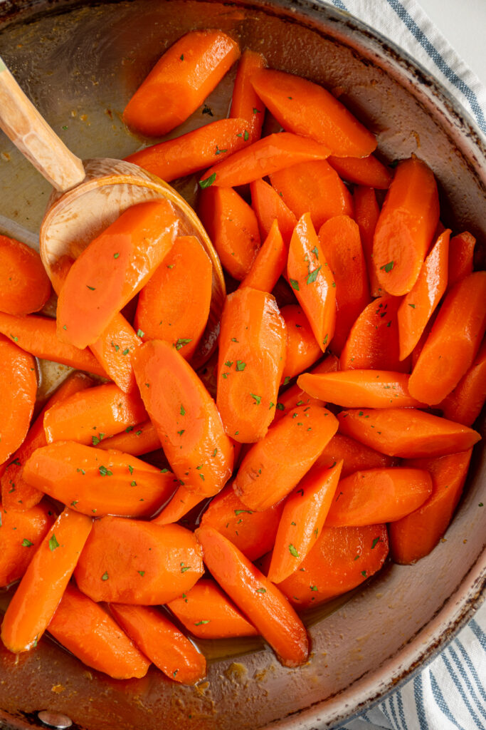 A skillet with honey glazed carrots with a wood spoon and garnished with a bit of fresh parsley.