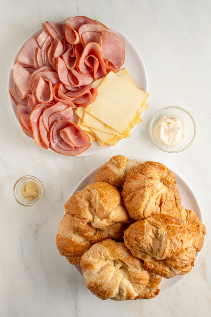Ingredients needed to make baked ham and cheese croissants laid out on plates on a countertop.