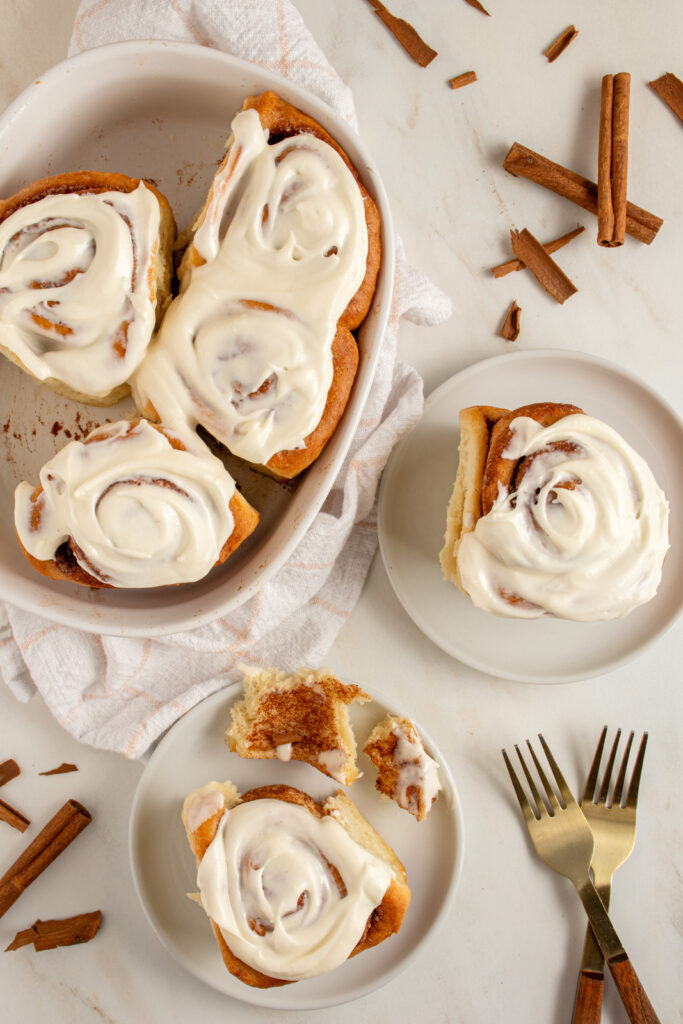 A pair of cinnamon rolls on small plates next to a baking dish filled with cinnamon rolls all covered in cream cheese frosting.