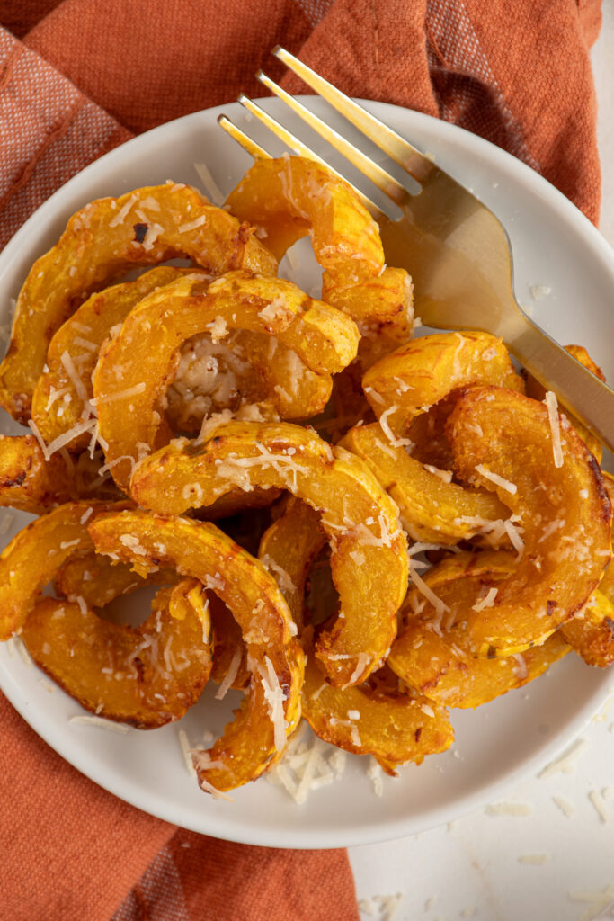 Delicata squash cooked golden brown with melted parmesan cheese on a white plate next to a fork.