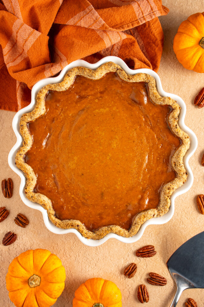 Overhead view of a white pie plate filled with pumpkin pie with a pecan infused crust surrounded by mini pumpkins and pecan halves.