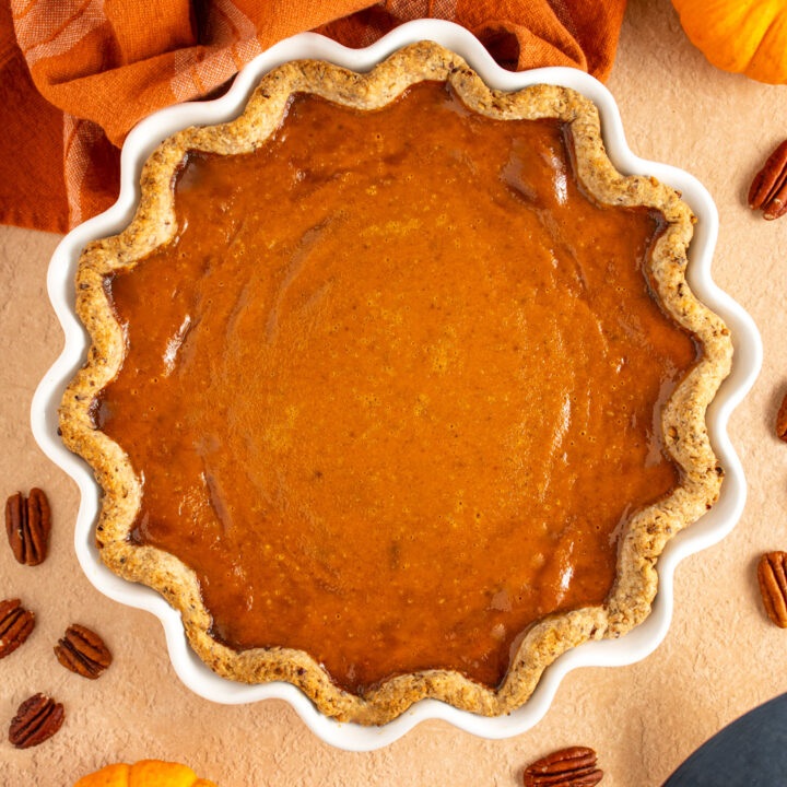 Overhead view of a white pie plate filled with pumpkin pie with a pecan infused crust surrounded by mini pumpkins and pecan halves.