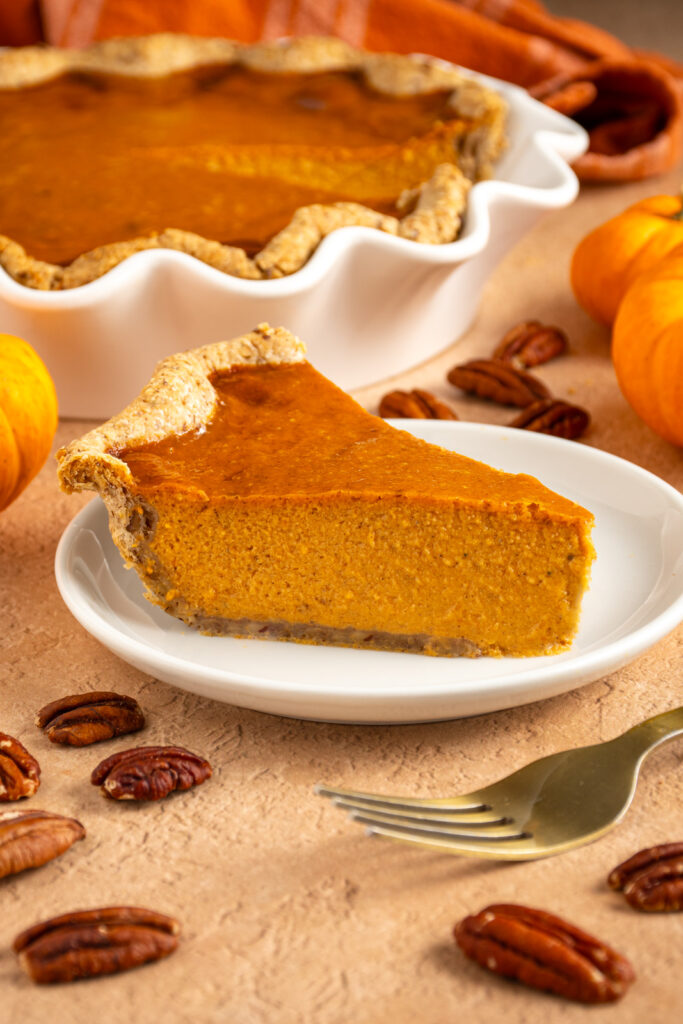 A slice od pumpkin pie with pecan crust on a small white plate surrounded by pecans and a pie plate filled with pumpkin pie.