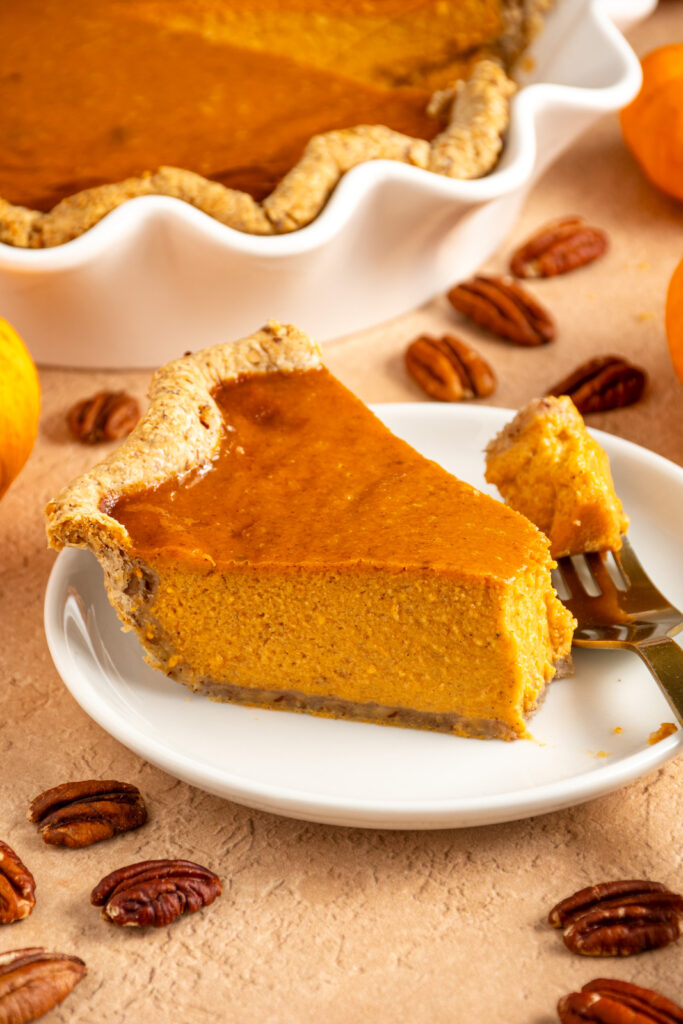 A fork with a piece of pumpkin pie with pecan crust and the slice of pie on a white plate showing off the orange pumpkin spiced filling.