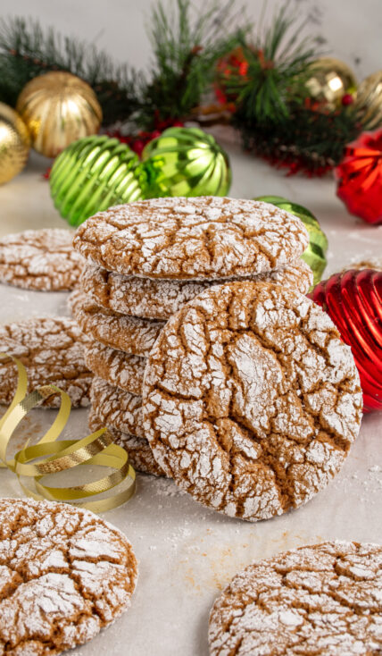A stack of gingerbread crinkle cookies on a sheet pan with one leaning against the stack surrounded by Christmas decorations.
