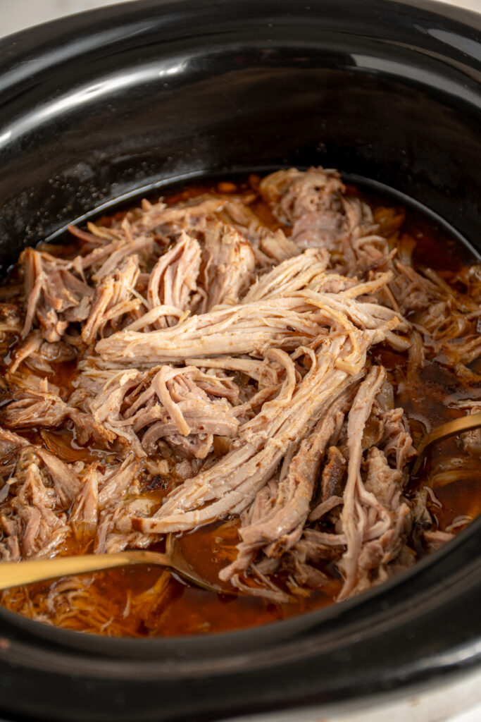 A black slow cooker with pulled pork and a serving spoon.