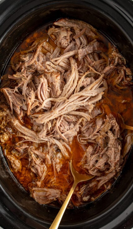Overhead view of a black crockpot filled with pulled pork with a gold serving spoon.