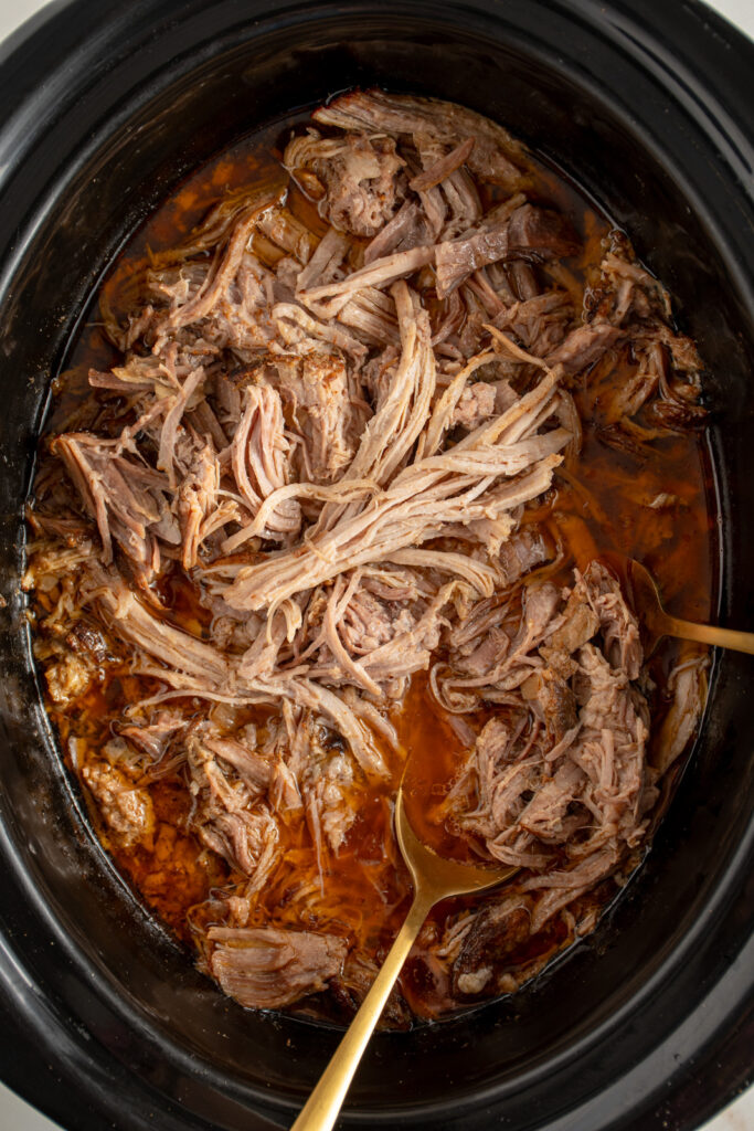 Overhead view of a black crockpot filled with pulled pork with a gold serving spoon.