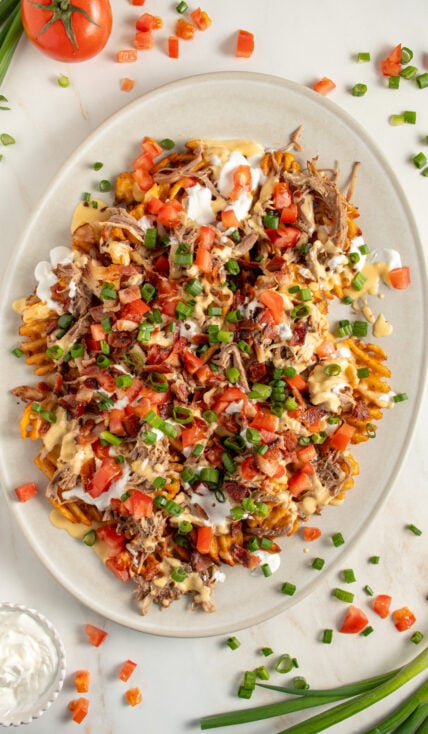 A large platter of pulled pork waffle fries piled with cheese sauce, sour cream, bacon, tomatoes, and green onions.