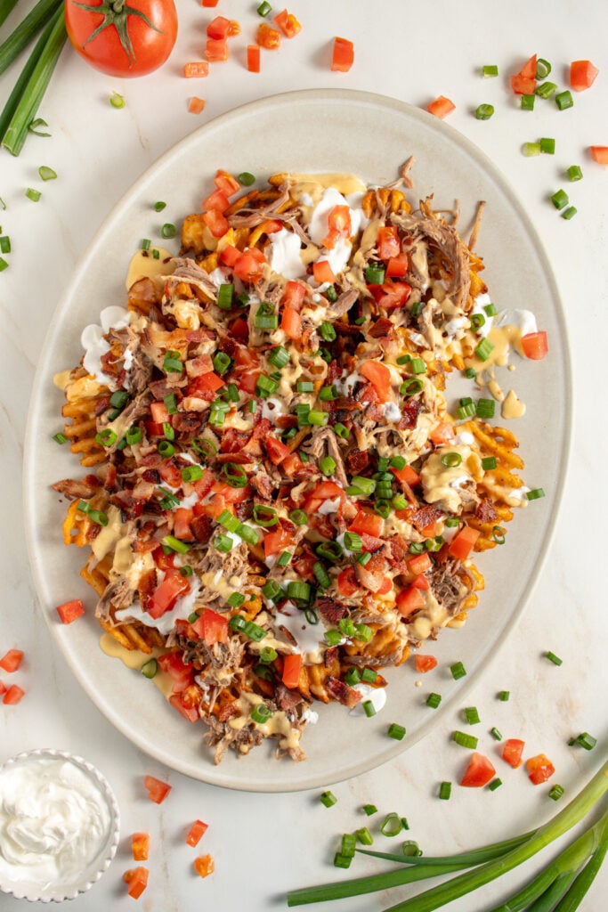A large platter of pulled pork waffle fries piled with cheese sauce, sour cream, bacon, tomatoes, and green onions.