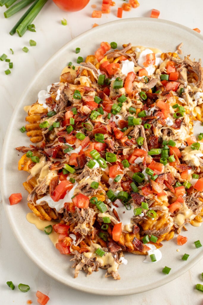 A large platter of pulled pork waffle fries piled with cheese sauce, sour cream, bacon, tomatoes, and green onions.