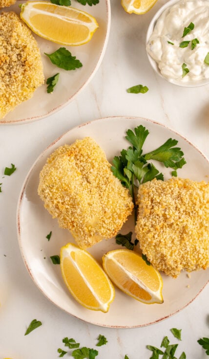 Plates with pieces of crispy baked breaded cod with lemons and fresh parsley.