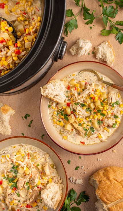 A brown bowl with chicken and corn chowder, a spoon, and a piece of crusty bread.
