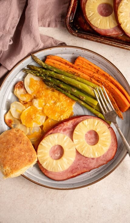 A plate with ham with pineapples, cheesy potatoes, asparagus, carrots, and biscuit.